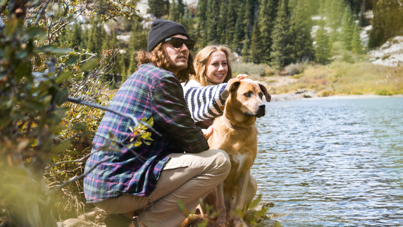 Two people and a dog sitting next to a lake.