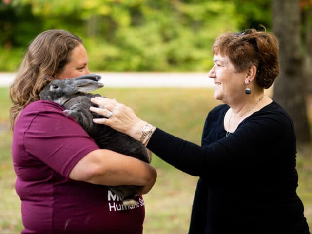 A woman holding a rabbit in her arms, alongside another woman petting the rabbit.