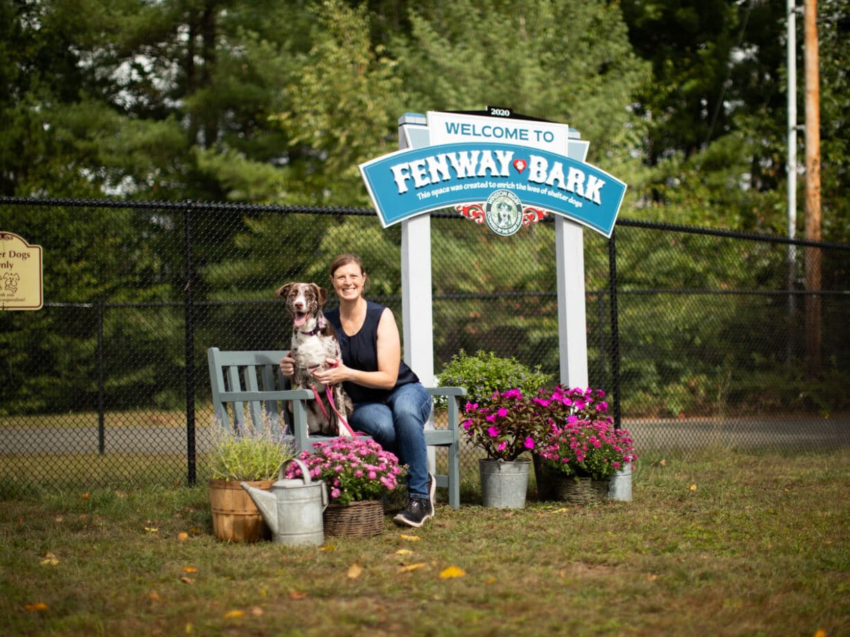A woman and a dog sitting on a bench in front of the Fenway Bark sign.