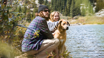 Two people and a dog sitting next to a lake.