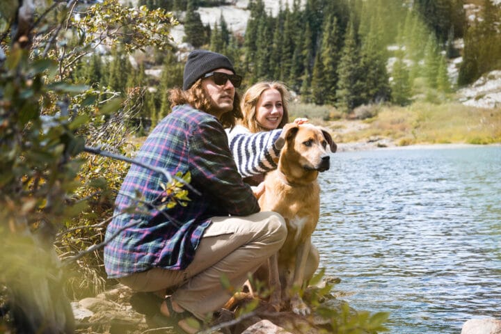 man and woman petting dog by riverbank.